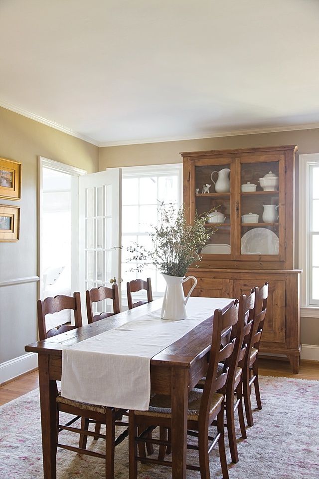 a dining room table with chairs and a vase on it's centerpiece in front of a china cabinet