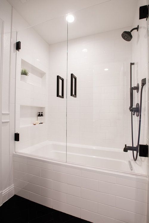 a white bathroom with black accents and tile flooring is pictured in this image, it has a glass shower door that opens up to reveal the bathtub area