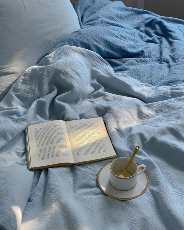 an open book on a bed with a cup of tea and saucer next to it