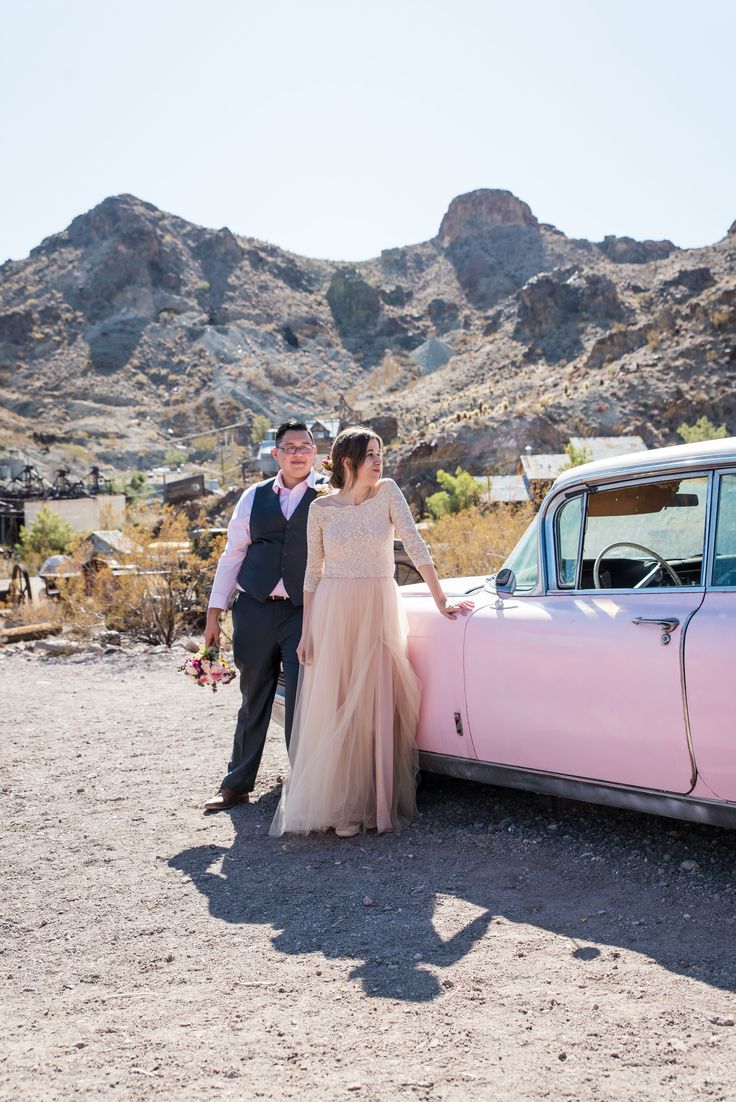 a man and woman standing next to an old pink car in the middle of nowhere