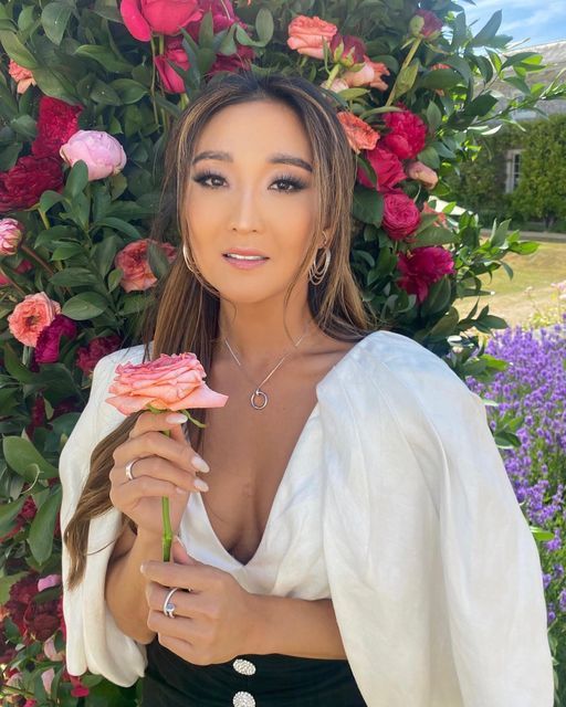 a beautiful woman holding a flower in front of some pink and red flowers on a bush