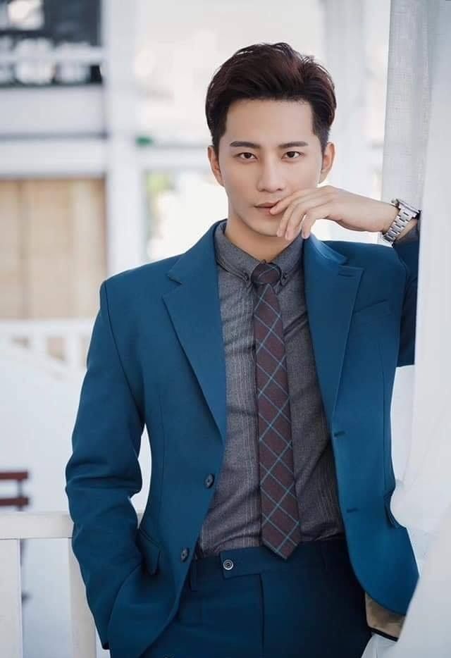 a man in a blue suit and tie leaning against a wall with his hand on his chin