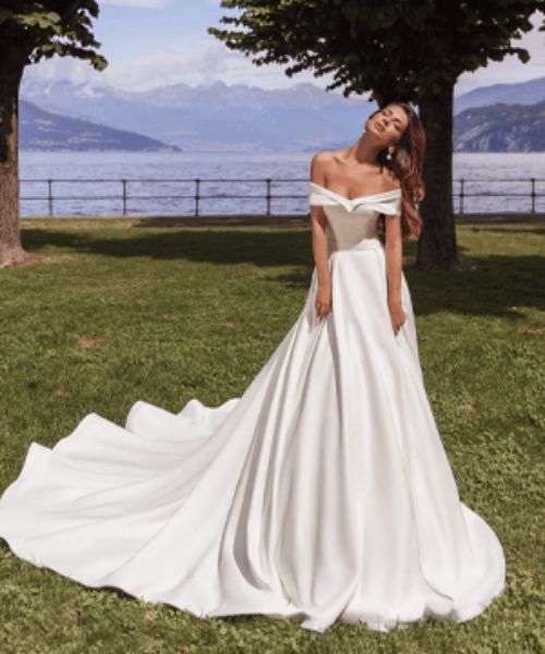 a woman in a white wedding dress standing on the grass near some trees and water