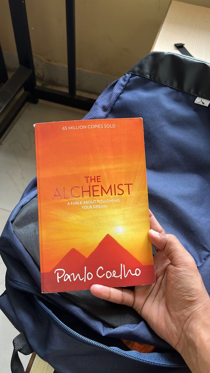 the alchemist book is in someone's hand on top of a backpack