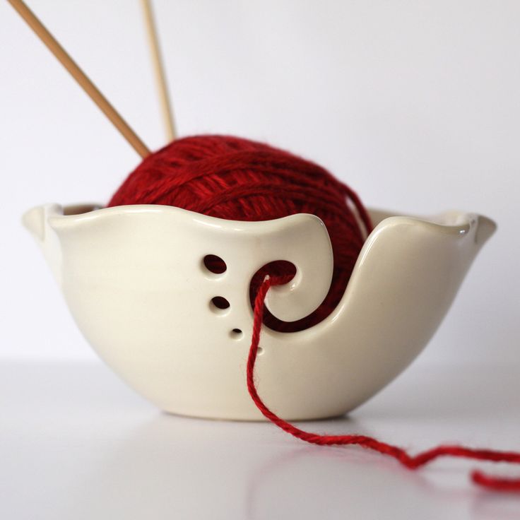 a red ball of yarn sitting in a white bowl next to two knitting needles and a crochet hook