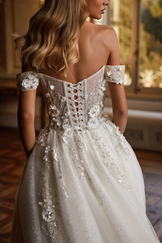 a woman in a white wedding dress with flowers on the back and shoulder, looking off to the side