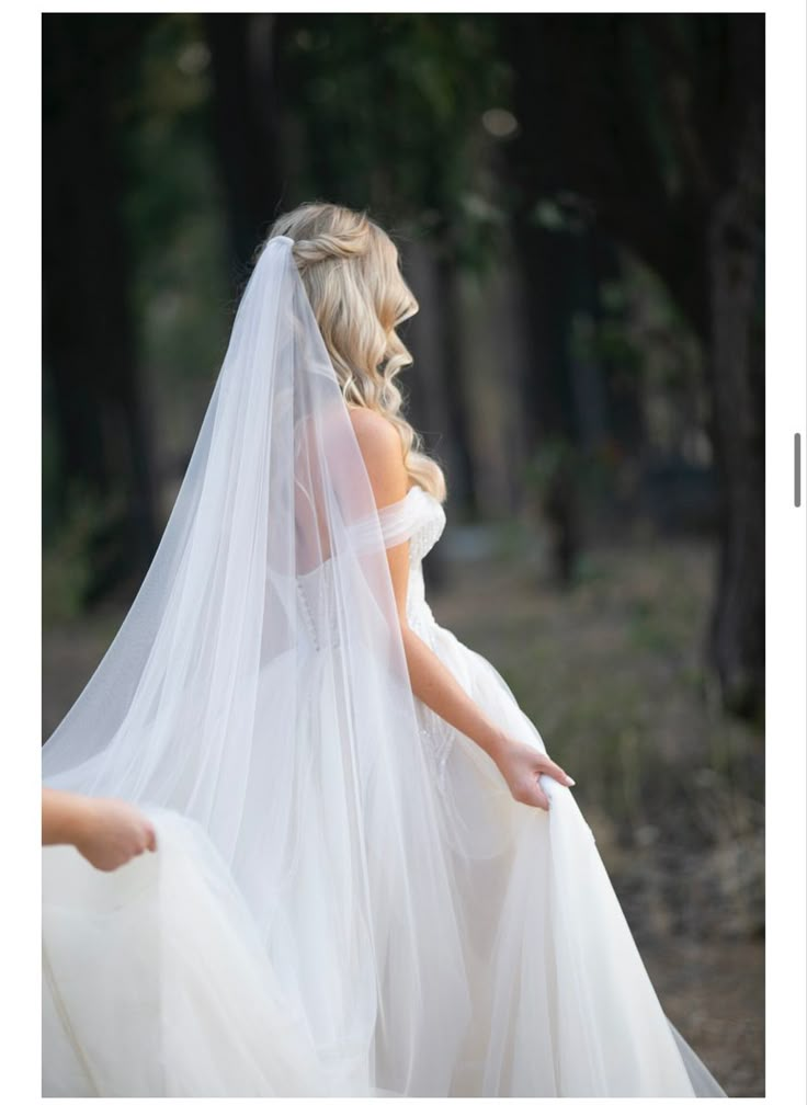 two brides in white dresses and veils walking through the woods with trees behind them