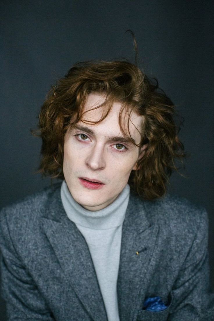 a young man with curly hair wearing a gray jacket and turtle neck sweater looking at the camera