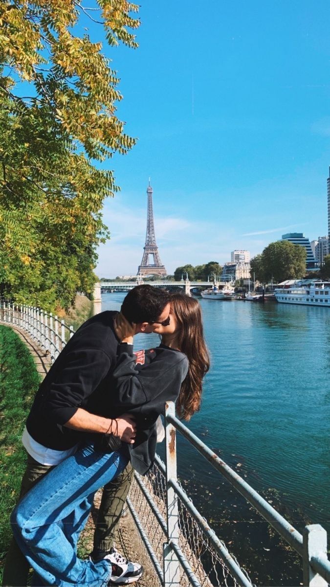 a man and woman leaning against a fence near the water in front of the eiffel tower