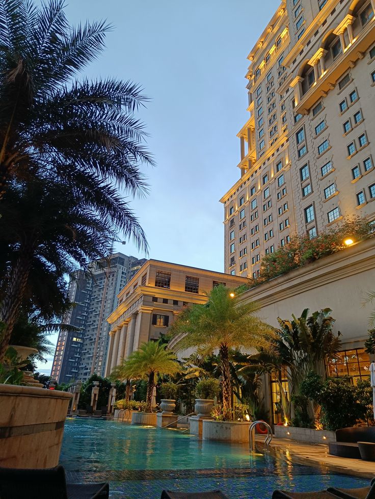 an outdoor swimming pool with chairs and palm trees in front of the hotel at dusk