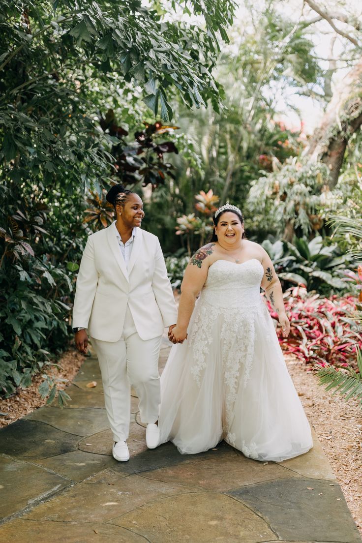 a bride and groom holding hands while walking down a path in front of tropical trees