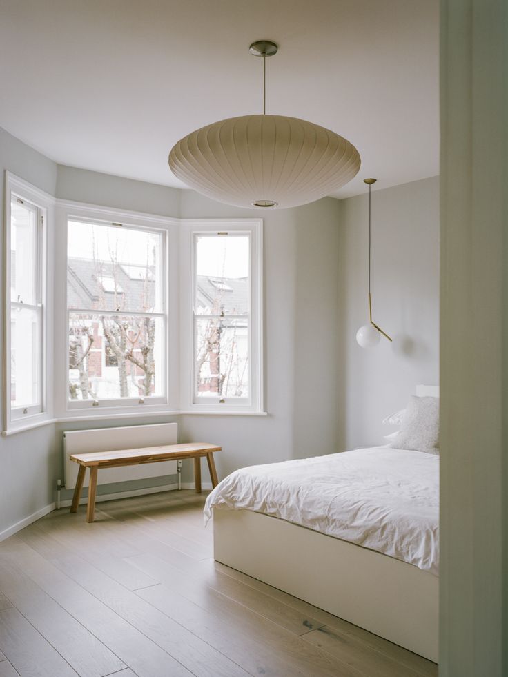 a white bed sitting under two windows in a bedroom next to a wooden table and bench