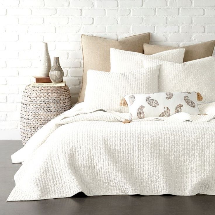 a bed with white sheets and pillows next to a basket on the floor in front of a brick wall