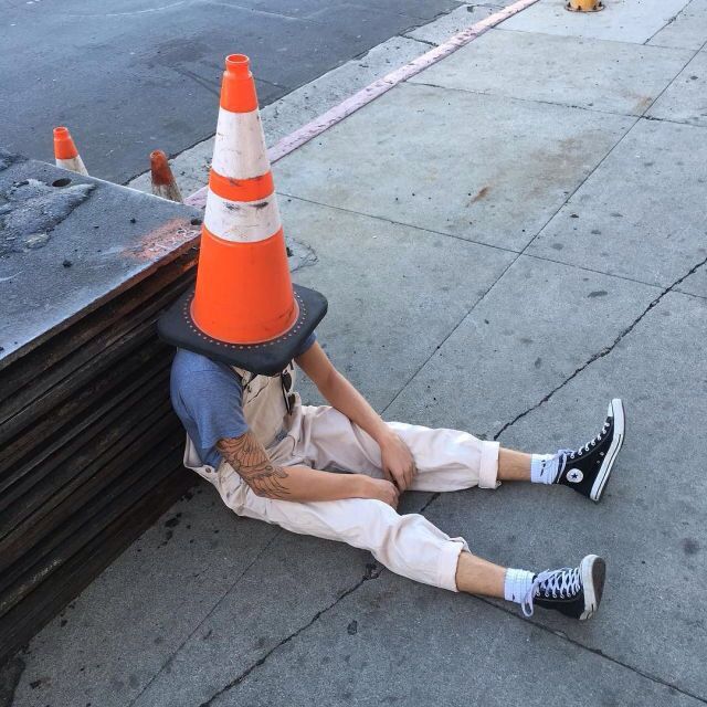 a person sitting on the sidewalk with an orange traffic cone over their head and feet