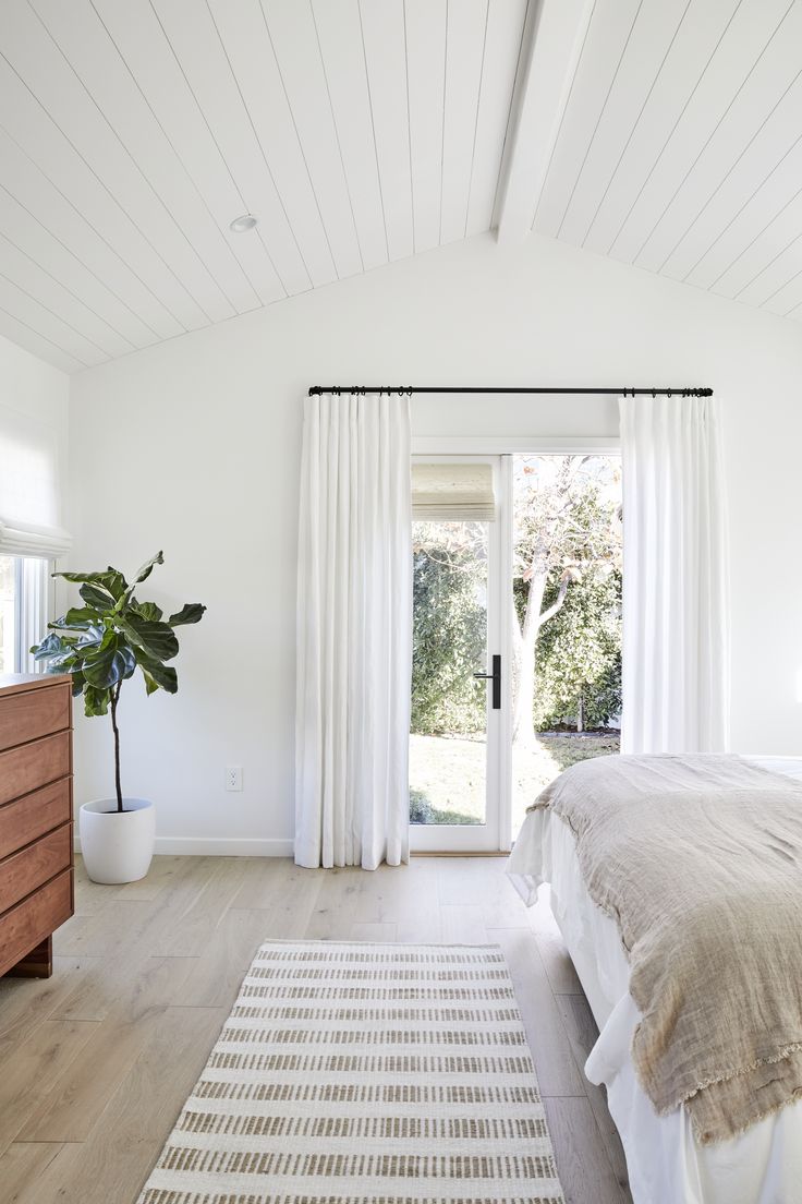 a bedroom with white walls and wood flooring has a large bed, wooden dresser, and sliding glass door that leads to an outdoor patio