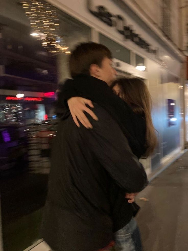 two people hugging each other in front of a building at night with lights reflecting off the windows