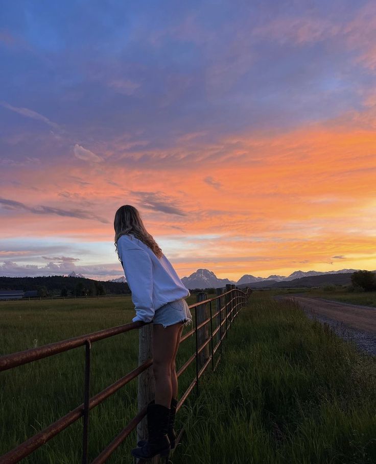 a woman leaning on a fence looking at the sunset over a field with mountains in the background