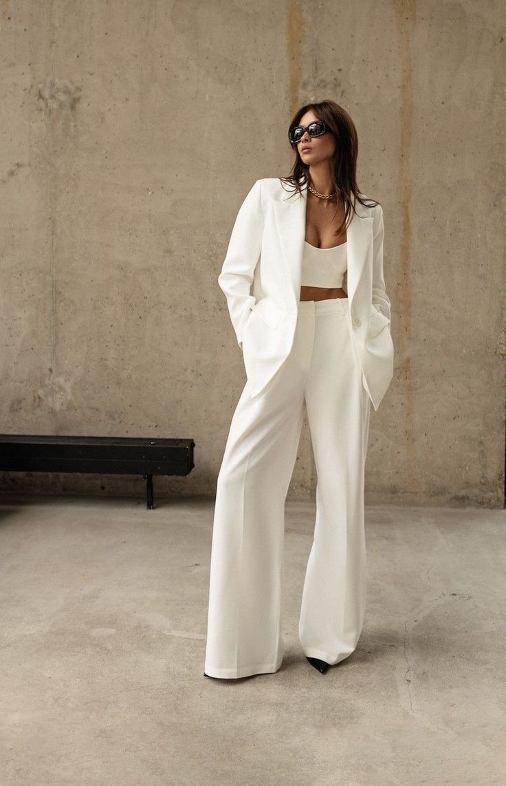 a woman standing in front of a concrete wall wearing white pants and a blazer