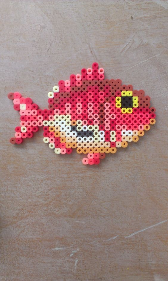 a red and yellow fish made out of perler beads on a table next to a cup