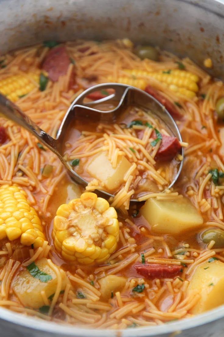 a pot filled with pasta and vegetables next to a ladle full of corn on the cob