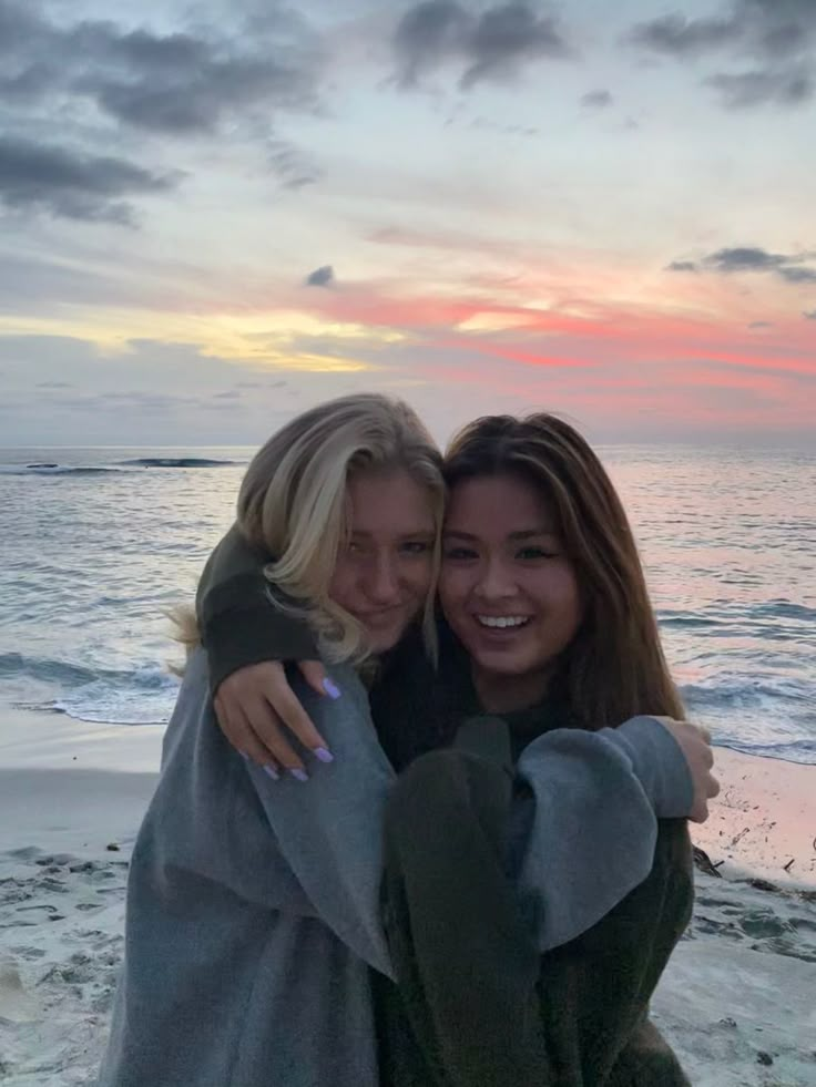 two young women hugging each other on the beach at sunset, with an ocean in the background