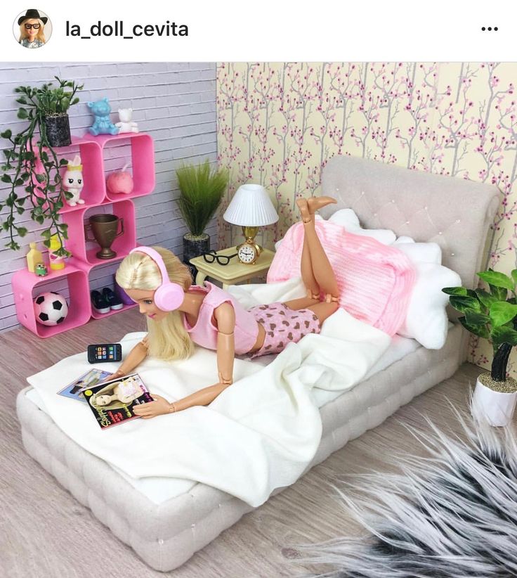 a barbie doll laying on top of a bed next to a lamp and bookshelf