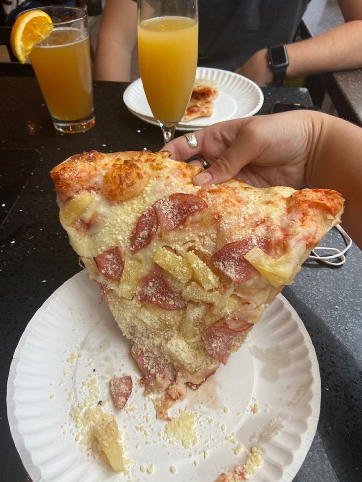 a slice of pizza on a paper plate with orange juice in front of it and two people sitting at a table