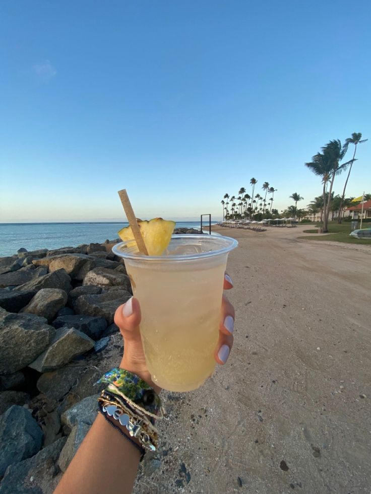 a person holding up a drink on the beach with a straw and an apple in it