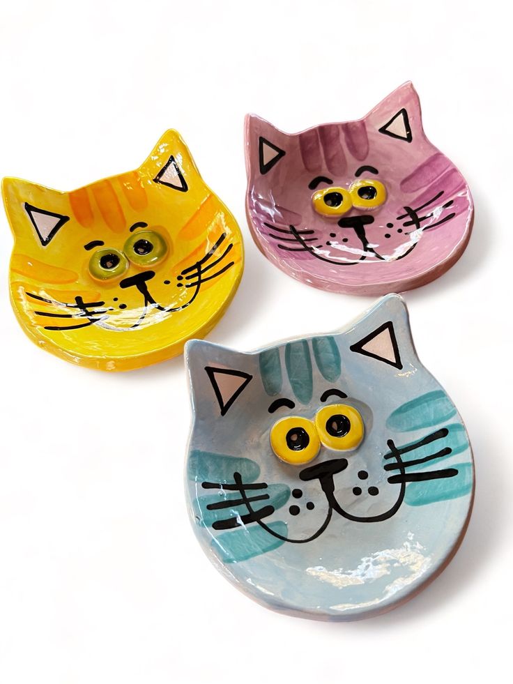 three cat shaped dishes with yellow eyes and one has a blue face, the other is pink