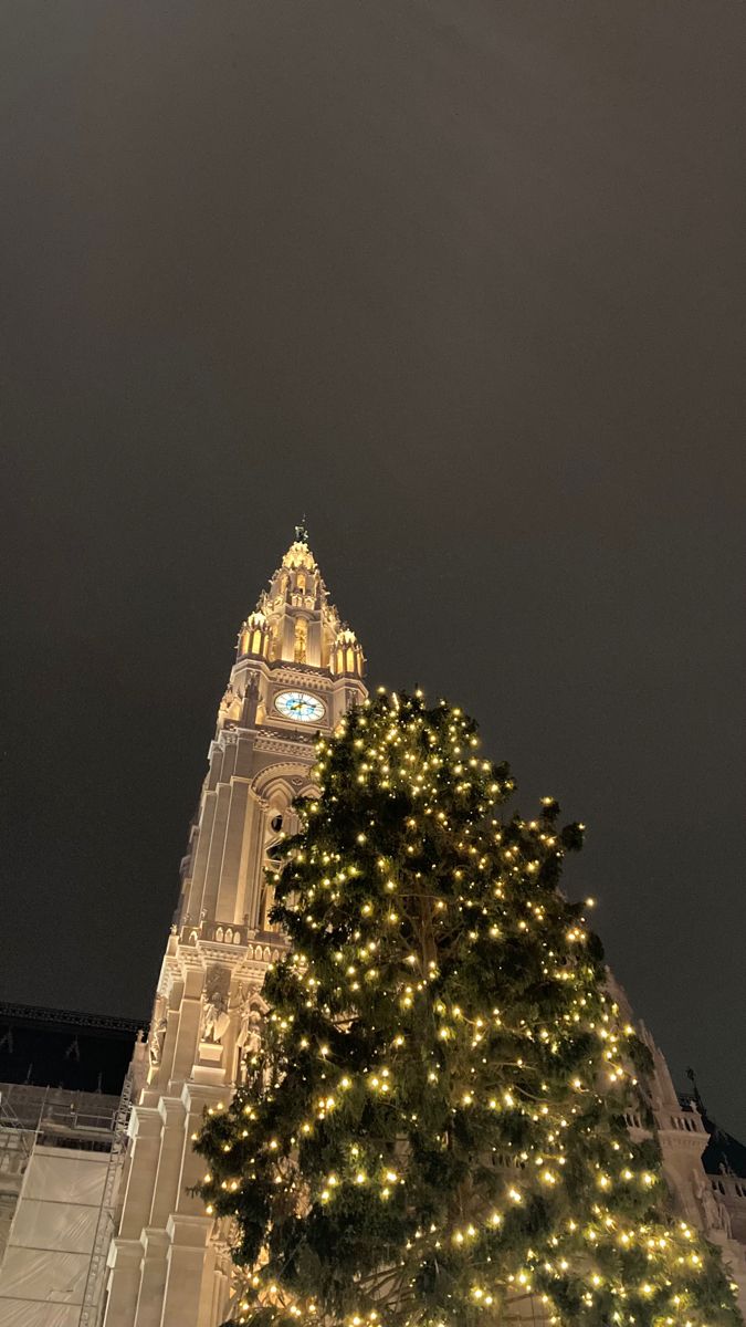 a very tall building with a clock on it's side and christmas lights around the tree