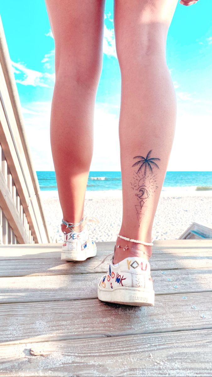 a woman's legs with a palm tree tattoo on her left leg, standing on a boardwalk