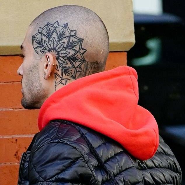 a bald man with a flower tattoo on his head looking off into the distance while standing next to a brick wall