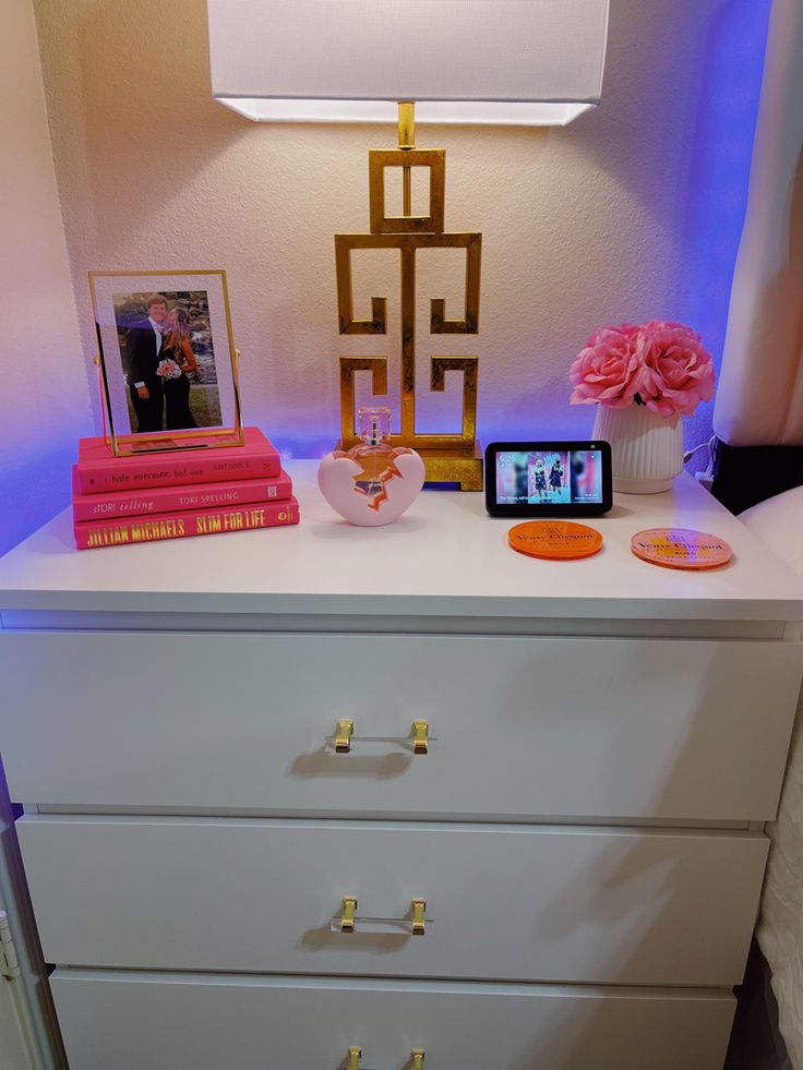 a white dresser with pink flowers and pictures on the top, next to a lamp