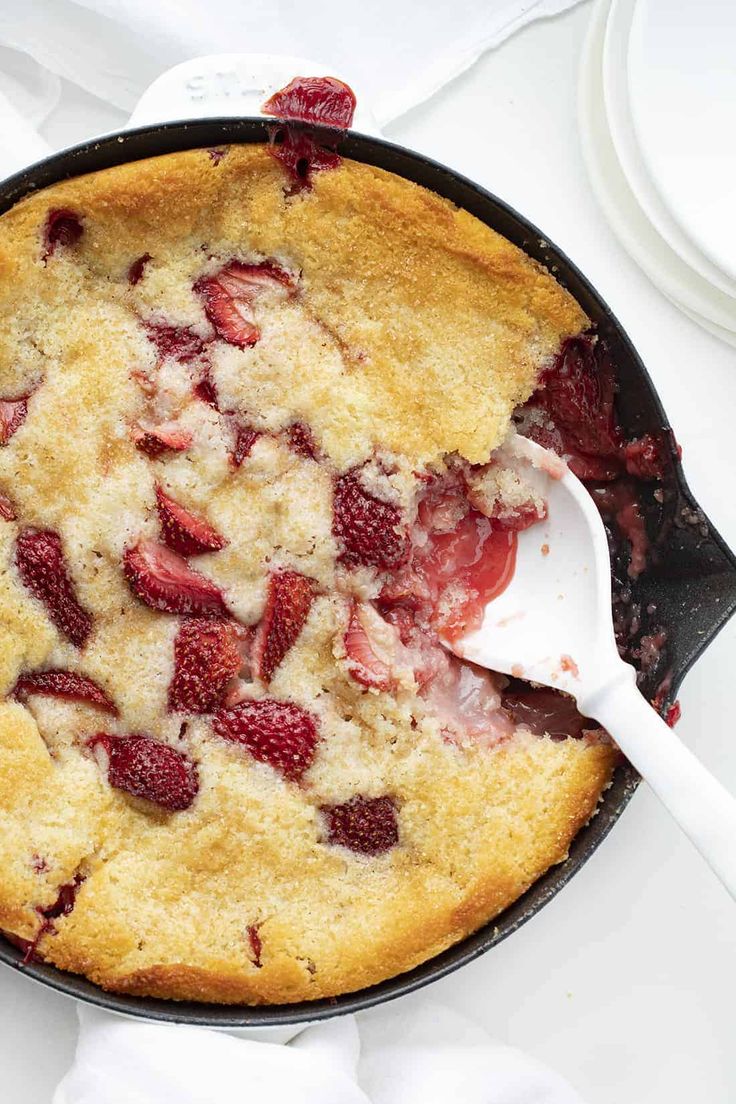 a skillet filled with strawberry cobbler on top of a white table next to plates and utensils