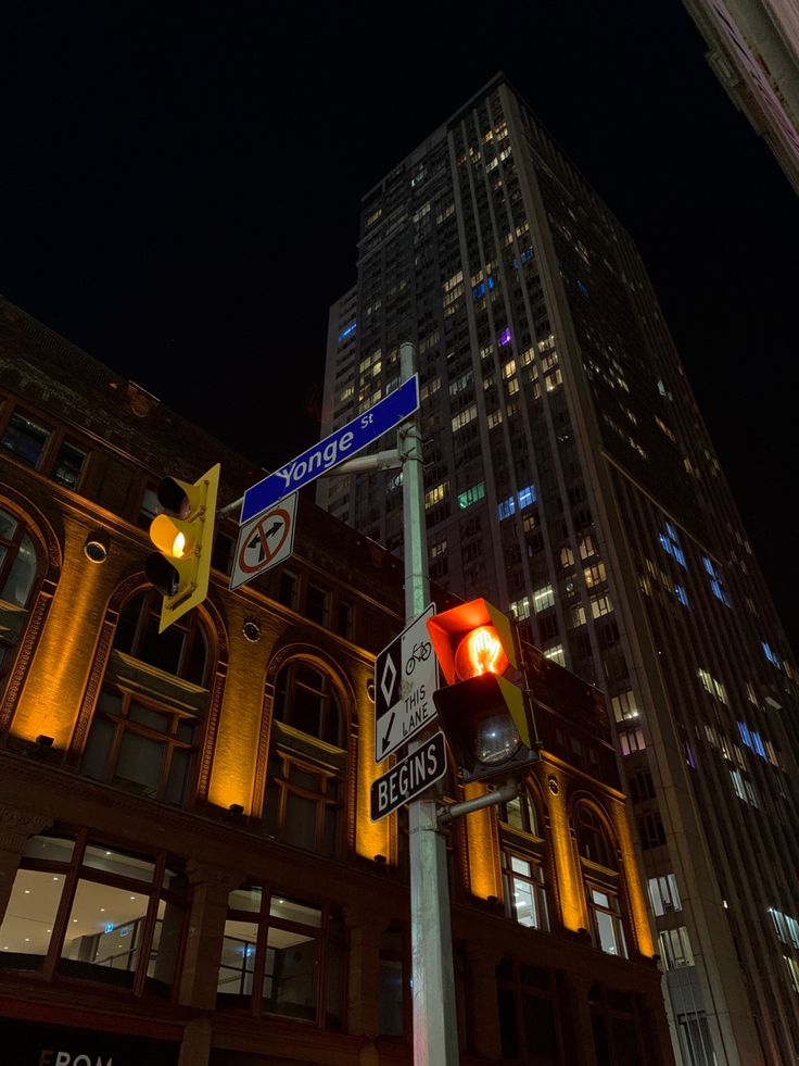 street signs and traffic lights in front of a tall building at night with buildings lit up