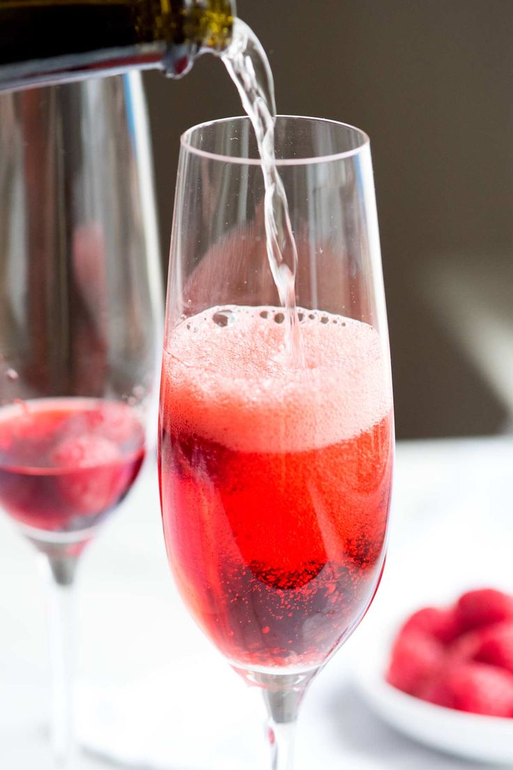 two wine glasses filled with red liquid being poured into one glass, and raspberries in the other