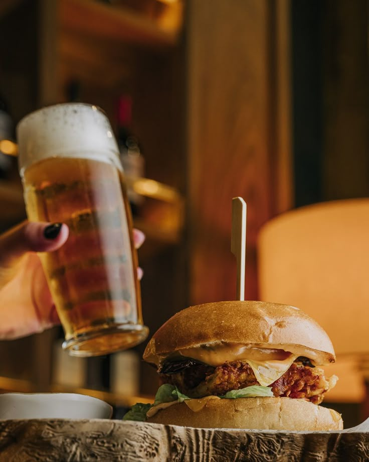 a person is holding a beer and a large hamburger on a wooden table in front of them