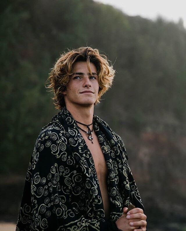 a man with no shirt on standing in front of some water and trees, wearing a shawl over his shoulders