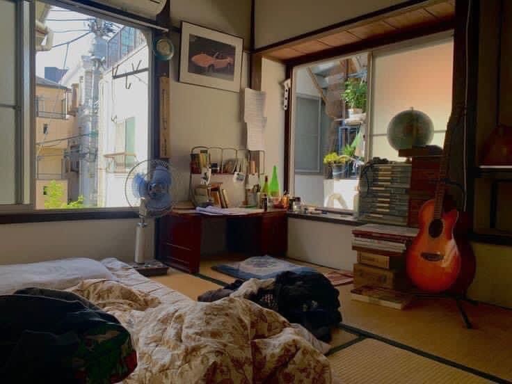 a bedroom with a guitar on the floor next to an open window and fan in front of it