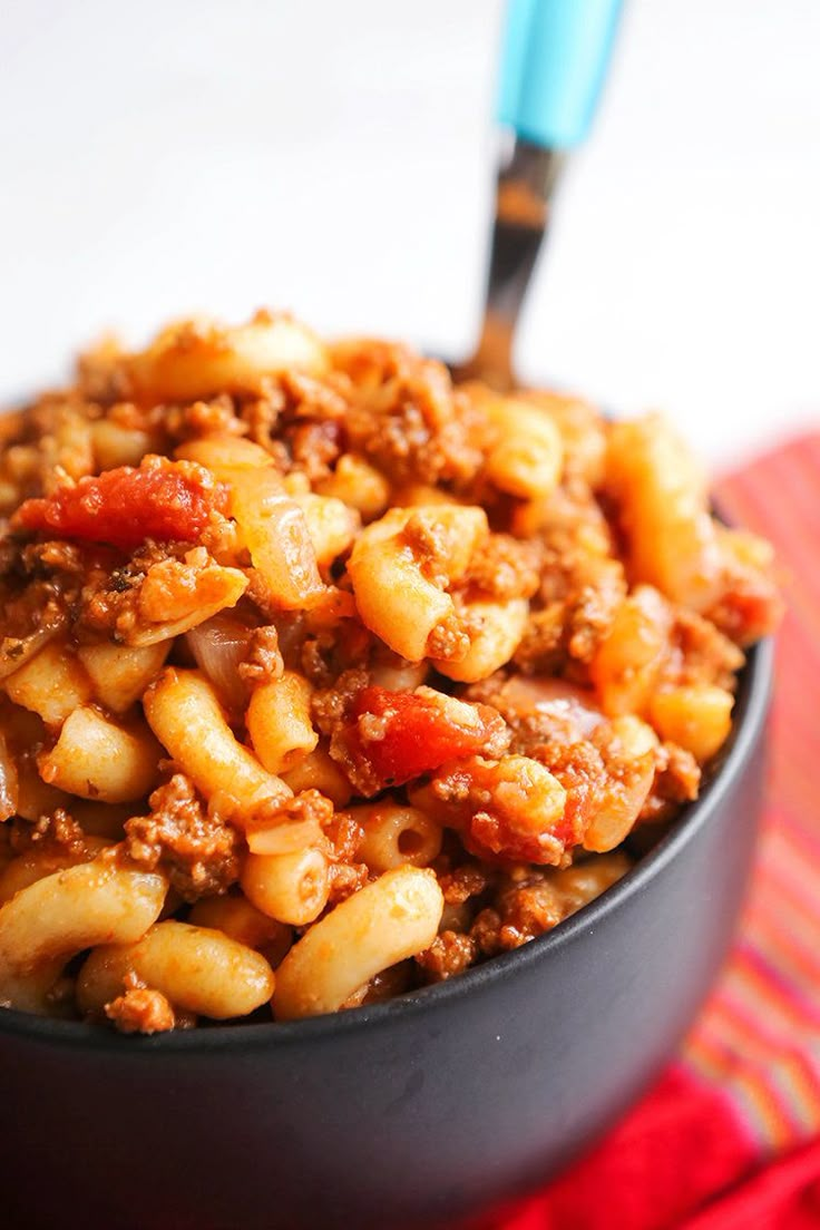 a black bowl filled with pasta and meat on top of a red cloth next to a blue fork