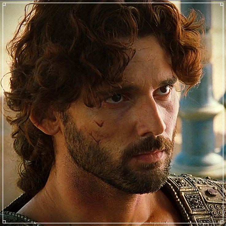 a close up of a man with long curly hair and beard wearing a roman costume