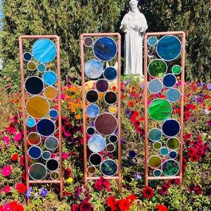 three colorful stained glass panels in front of a statue and flower garden with flowers around them