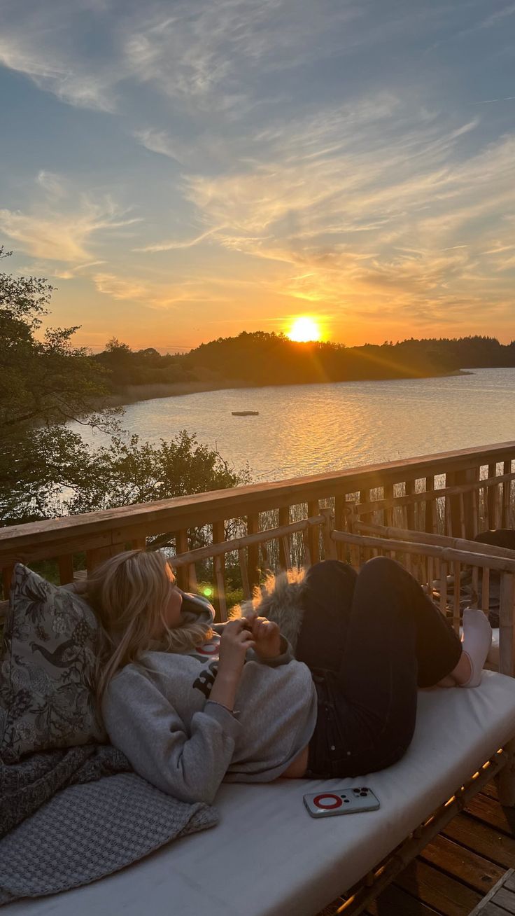 two people laying on a deck with the sun setting in the background and water behind them