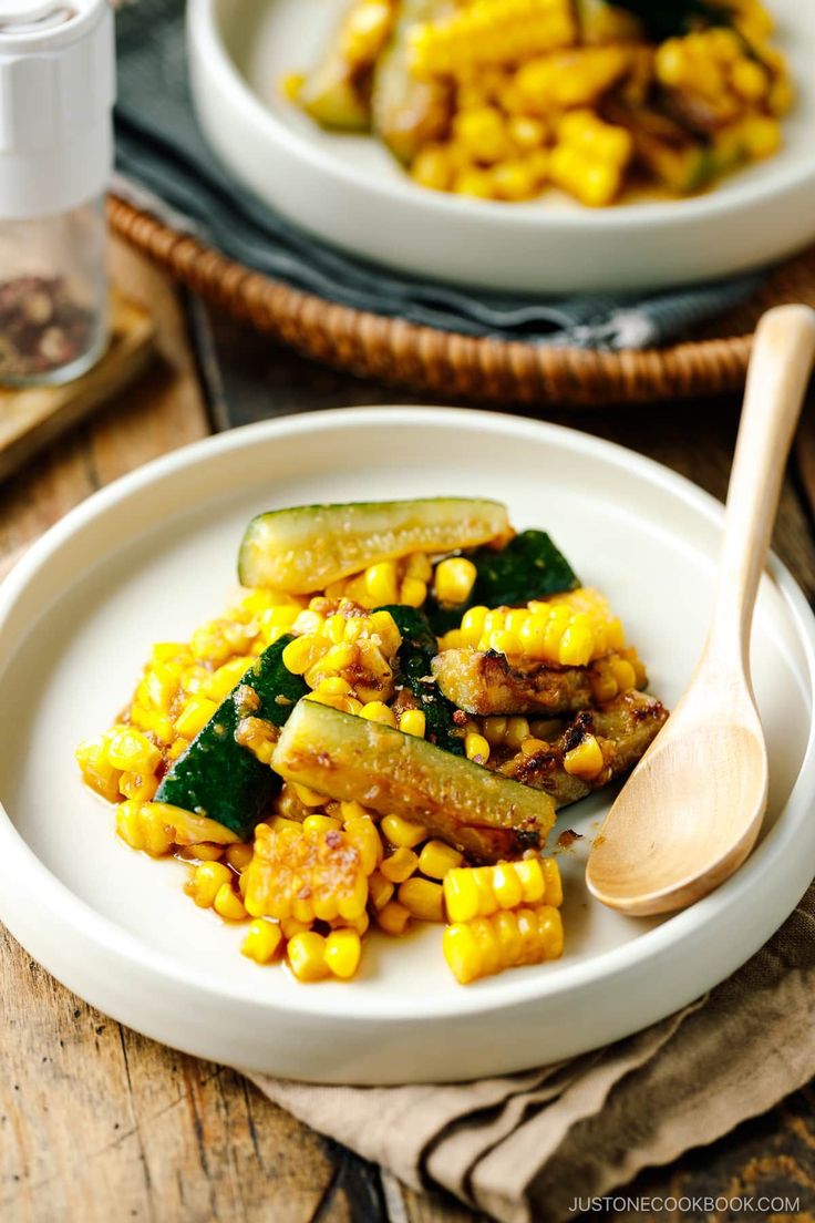 two white plates filled with corn and zucchini on top of a wooden table