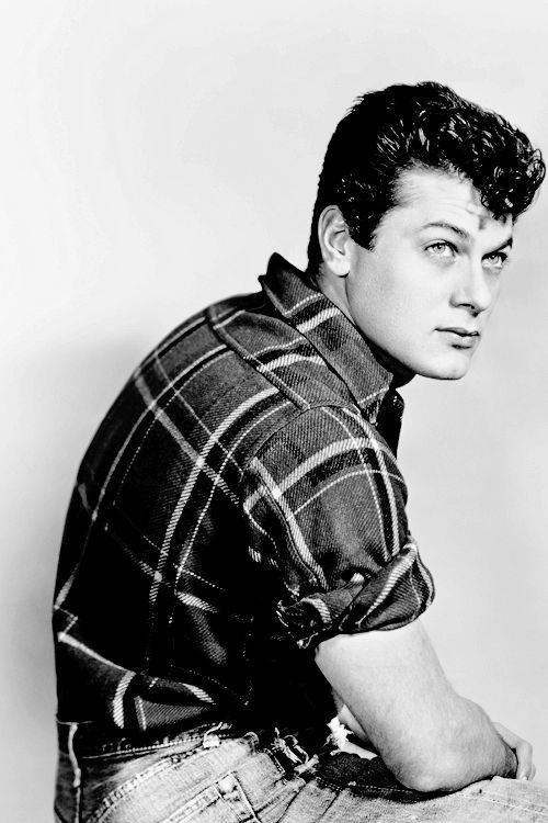 a black and white photo of a man in plaid shirt leaning on a stool with his hand on his hip