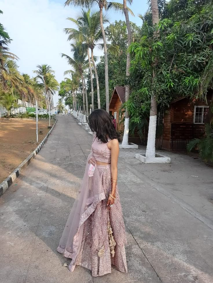 a woman in a long dress walking down the street with palm trees behind her and a small hut on the other side