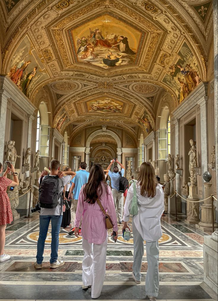 several people are walking in an ornate hallway with paintings on the walls and ceiling above them