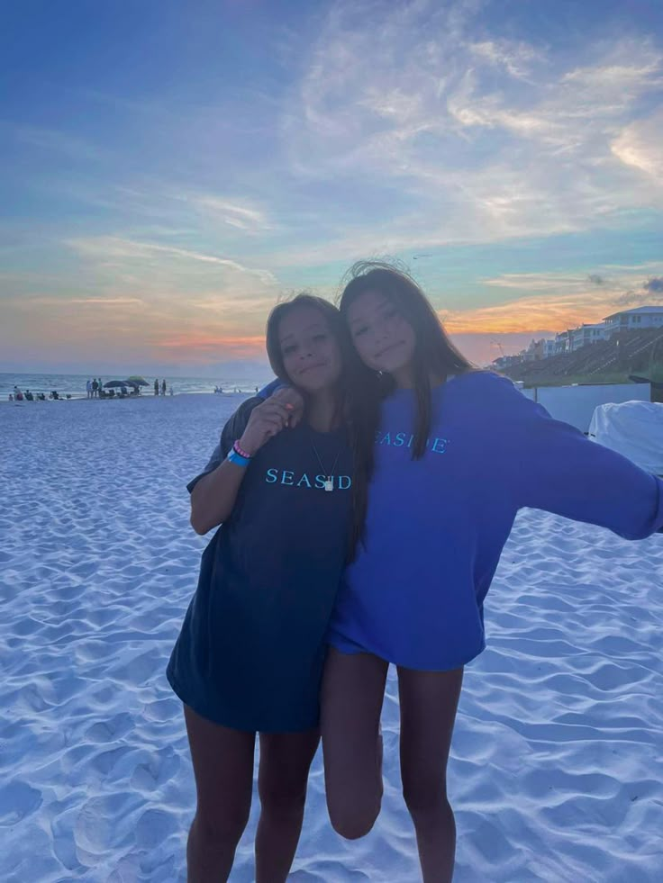 two girls standing in the sand with their arms around each other