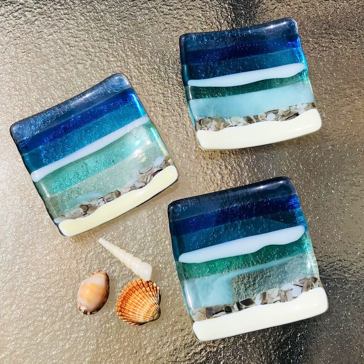 three square glass dishes sitting on top of a metal surface next to sea shells and seashells