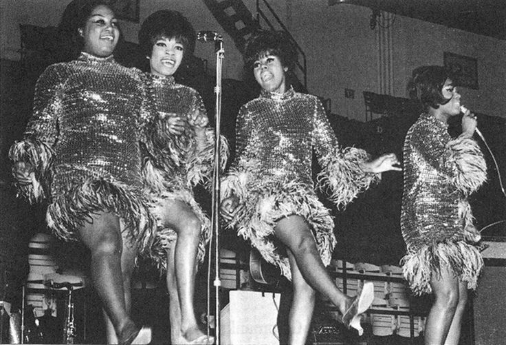 three women in sequin dresses singing on stage with microphone and two men standing behind them